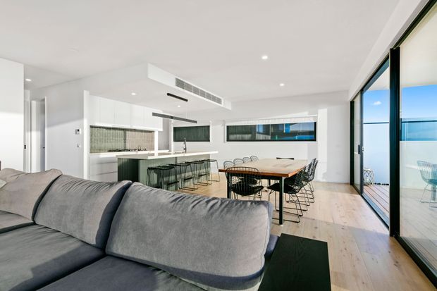 Bright open-plan contemporary living, dining and kitchen area with a large grey sectional sofa in the foreground, a white streamlined kitchen with island and bar seating, and a timber dining table with wire-frame chairs. Full-height black-framed glazing opens toward an outdoor deck, while pale timber flooring and white surfaces create an airy, minimal interior.