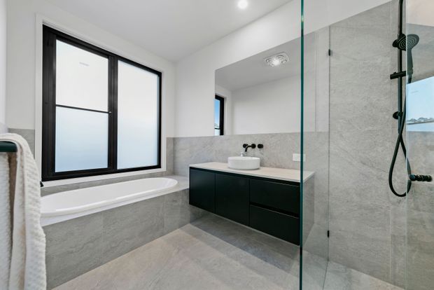 Contemporary bathroom with a freestanding bathtub beneath black-framed frosted windows, a floating dark vanity with a white basin, and a large frameless glass walk-in shower. Light grey stone-look wall and floor finishes create a clean, restrained palette.