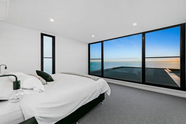 Bright minimalist bedroom with a low-profile bed dressed in white linens, dark green accents, white walls and ceiling, and a grey carpeted floor. Full-height black-framed glazing spans one wall, overlooking a calm coastal horizon and an expansive rooftop or deck-like surface.