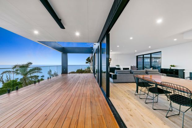 A contemporary open-plan living and dining area opening onto a large timber deck with glass balustrades and expansive ocean views. The interior has white walls and ceilings, light timber flooring, black-framed glazing, a grey sectional sofa, and a black wire-frame dining set. A covered outdoor section with dark structural beams extends over the deck, creating a strong indoor–outdoor connection.