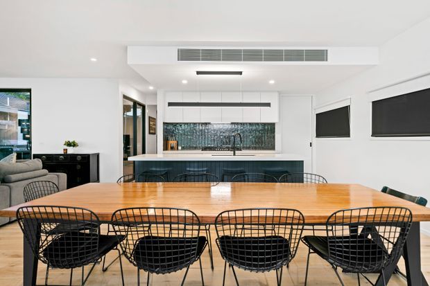 Bright open-plan kitchen and dining area with a large timber dining table surrounded by black wire-frame chairs. The kitchen features white cabinetry, a dark stone or tiled splashback, a dark island bench, integrated appliances, black-framed glazing, and roller blinds. A living area is partially visible at left.