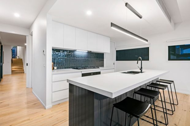 Bright contemporary kitchen with white full-height cabinetry, a large white island with integrated sink and black vertical-panelled base, black bar stools, linear pendant lighting, and a reflective blue-grey tiled splashback. Pale timber flooring continues into the adjacent hallway, which leads to a short staircase.