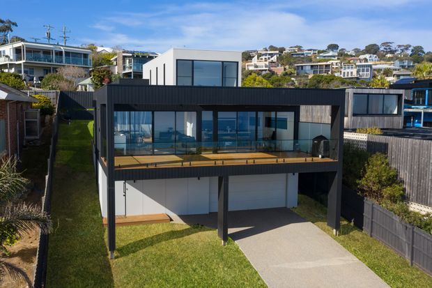 Aerial exterior view of a contemporary multi-level residence on a sloping suburban site. The home features a dark vertically clad upper volume, extensive floor-to-ceiling glazing, a broad timber deck, white lower-level walls, and an open ground-level carport. Neighbouring hillside homes and landscaped gardens are visible in the background.