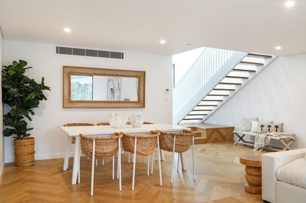 Bright open-plan dining and stair area with a long white dining table, woven rattan dining chairs, timber herringbone flooring, white walls, a large framed mirror, indoor plants, and a contemporary staircase with a white vertical balustrade and timber treads. A portion of the living area is visible at right.