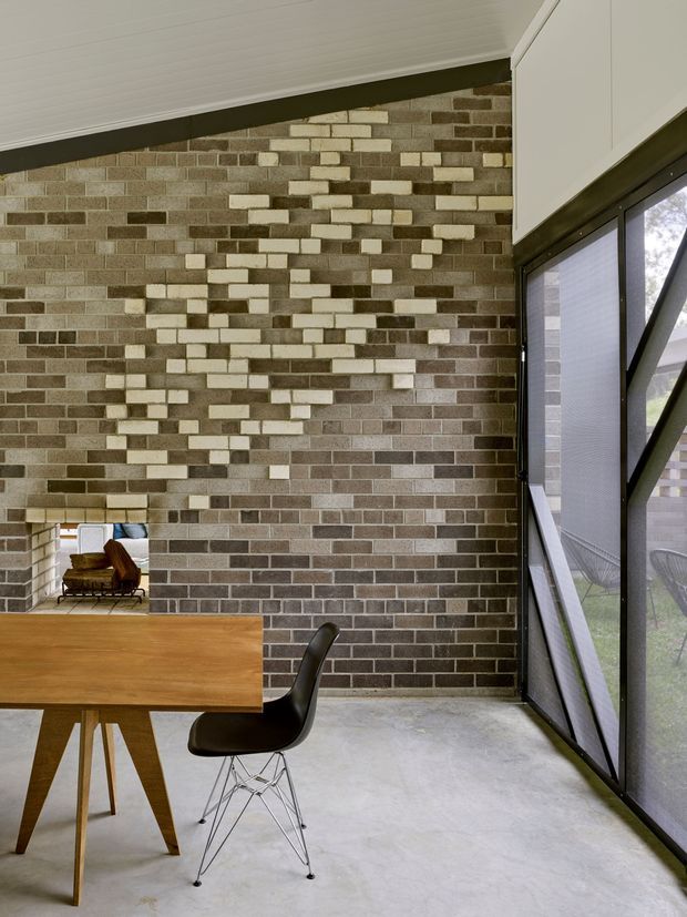 A contemporary dining area with a timber dining table and black chair beside a full-height glazed wall. The room features an exposed brick feature wall with a varied pattern of light and dark bricks, a pale white-lined sloped ceiling, and a polished concrete floor. A secondary seating area is visible through an opening at left.