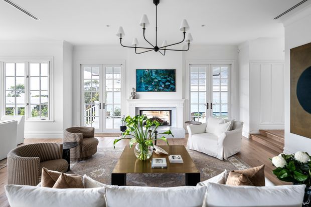 Bright, refined living room with white panelled walls, French doors and windows, a central fireplace, dark sculptural chandelier, neutral upholstered seating, patterned rug, and a timber coffee table with plants. A short set of steps leads to an adjacent level, while artwork and flowers add restrained colour.
