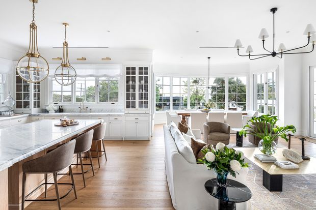 Bright, open-plan Hamptons-style kitchen and living area with extensive white cabinetry, a large marble island, pendant lights, timber flooring, and abundant floor-to-ceiling glazing. The living zone includes a white sofa, armchairs, a black coffee table, indoor plants, and a patterned rug, creating an airy, refined and welcoming interior.