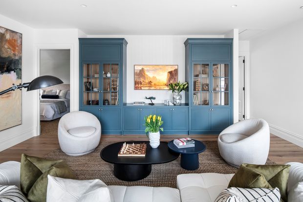 Bright, refined living room with a white sectional sofa in the foreground, sculptural white lounge chairs, black nesting coffee tables, and a large blue built-in cabinet wall with glazed display shelving. White walls and ceiling, timber flooring, a natural woven rug, artwork, and indoor plants create a contemporary yet classic interior.