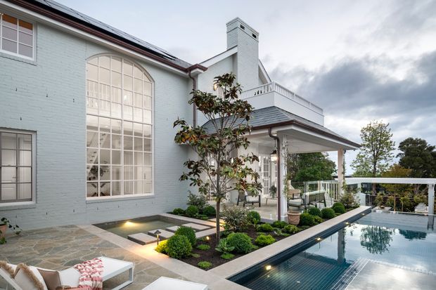 A refined residential courtyard and pool area beside a pale blue rendered brick home. The space features a large reflective swimming pool, stone-paved terrace, landscaped garden beds with clipped shrubs and a small tree, and a covered veranda with white columns and balustrading. The house has a dark pitched roof, prominent arched multi-pane window, and a tall chimney, creating a traditional architectural character with contemporary outdoor detailing.