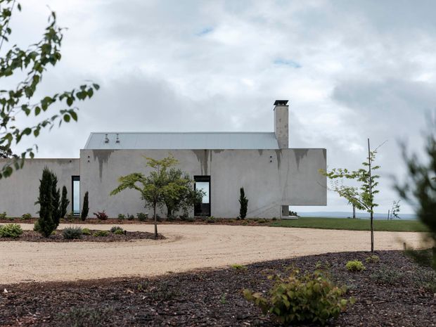 A minimalist single-storey concrete house with a low rectangular form, flat roof, and prominent chimney. The weathered pale grey facade has narrow dark-framed openings and a projecting side volume. It sits in a sparsely landscaped gravel setting with young trees and distant water visible beyond the property.