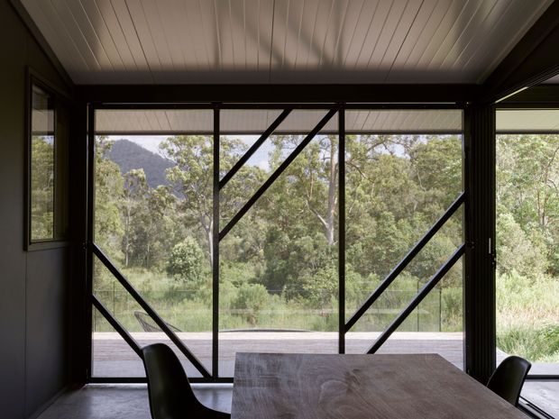 A dark, contemporary dining area framed by expansive floor-to-ceiling glazing with bold black diagonal structural bracing. A timber dining table and black chairs sit beneath a white linear-panel ceiling, opening onto a timber deck and a leafy, mountainous landscape.