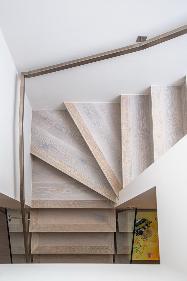 A close-up view looking up at a compact, sculptural staircase with pale timber-clad treads and risers arranged around a central turning flight. Slim dark metal handrails and supports frame the stairwell, while white walls and ceiling create a clean, bright setting. A colourful artwork is partially visible below the stairs.