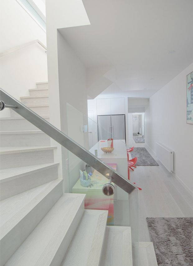 Bright minimalist interior view from a stairway, showing pale timber or wood-look stair treads, a glass balustrade with brushed metal handrail, white walls and ceiling, and an open-plan dining area beyond. The dining space contains a white table with red chairs, while a grey rug and hallway are visible in the background. A skylight or high-level glazing brings in abundant natural light.