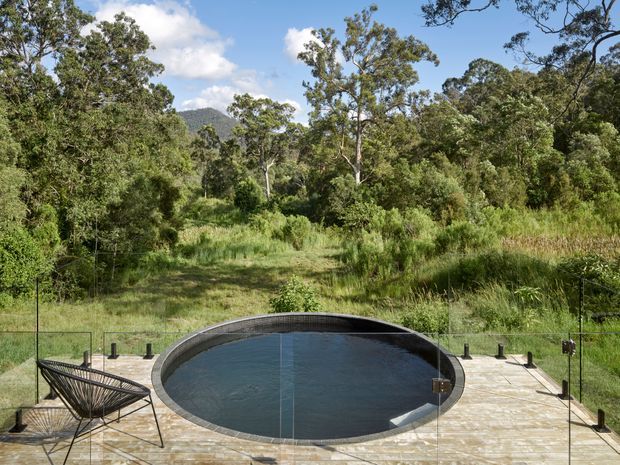 A landscaped outdoor pool area with a dark oval swimming pool set into a large pale stone or concrete-paved terrace. Slim black glass or mesh safety fencing surrounds the pool, with a sculptural black wire lounge chair at the edge. Dense native-looking vegetation and wooded hills form the background under a bright blue sky.