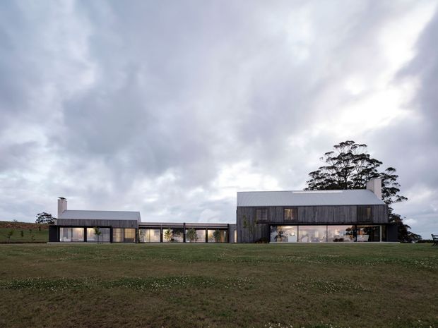 A low, elongated contemporary residence set in an expansive grassy rural landscape. The building comprises several connected single-storey volumes with dark vertical cladding, broad glazed openings, and simple metal roof planes. The overcast sky and mature trees create a subdued, minimalist architectural setting.