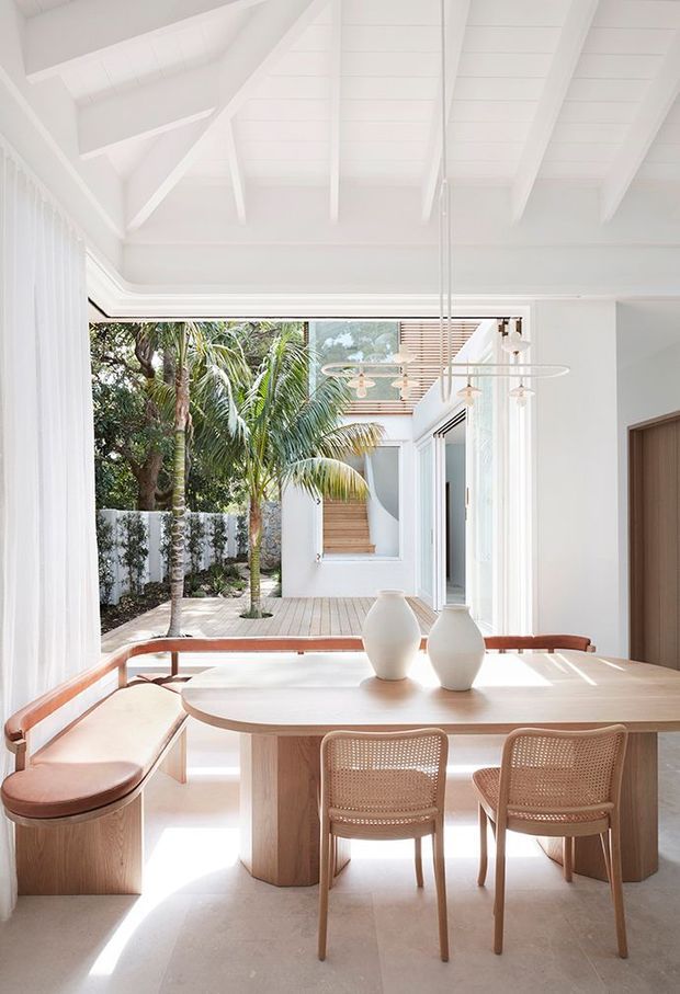 A bright, refined dining area with a large rounded timber dining table, built-in curved bench seating, and woven-back dining chairs. White walls and exposed white-painted rafters create an airy high-ceilinged interior, opening directly onto a sunlit timber deck and tropical courtyard. Contemporary pendant lighting and simple ceramic vessels complete the calm, natural scheme.