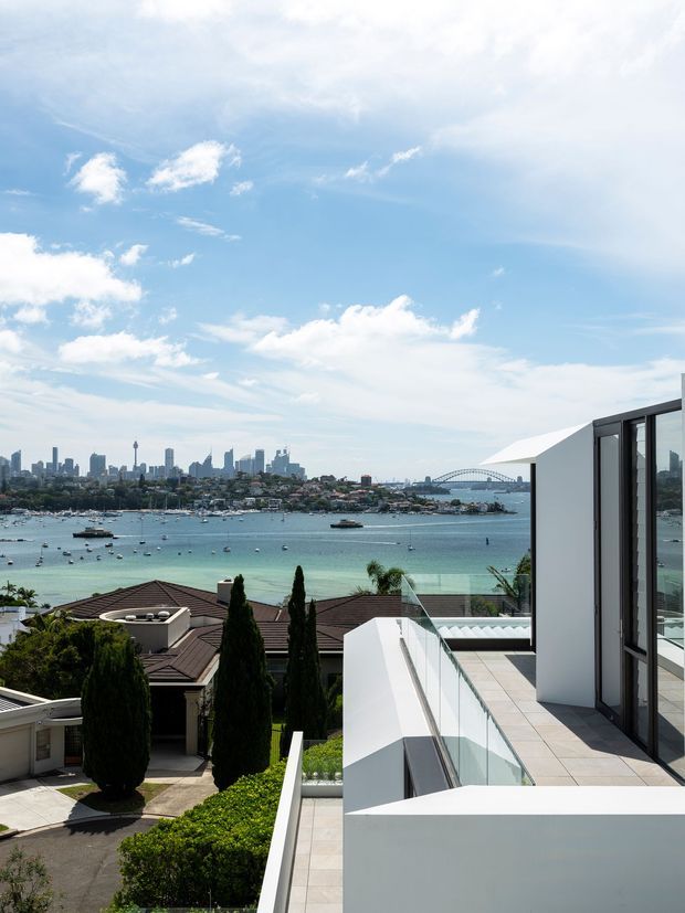 A bright exterior architectural view from a contemporary terrace overlooking a broad harbour, with a dense city skyline and a large suspension bridge in the distance. The foreground shows crisp white rendered walls, pale tiled terrace paving, frameless glass balustrades, dark-framed glazing, landscaped hedges and neighbouring residential rooftops.