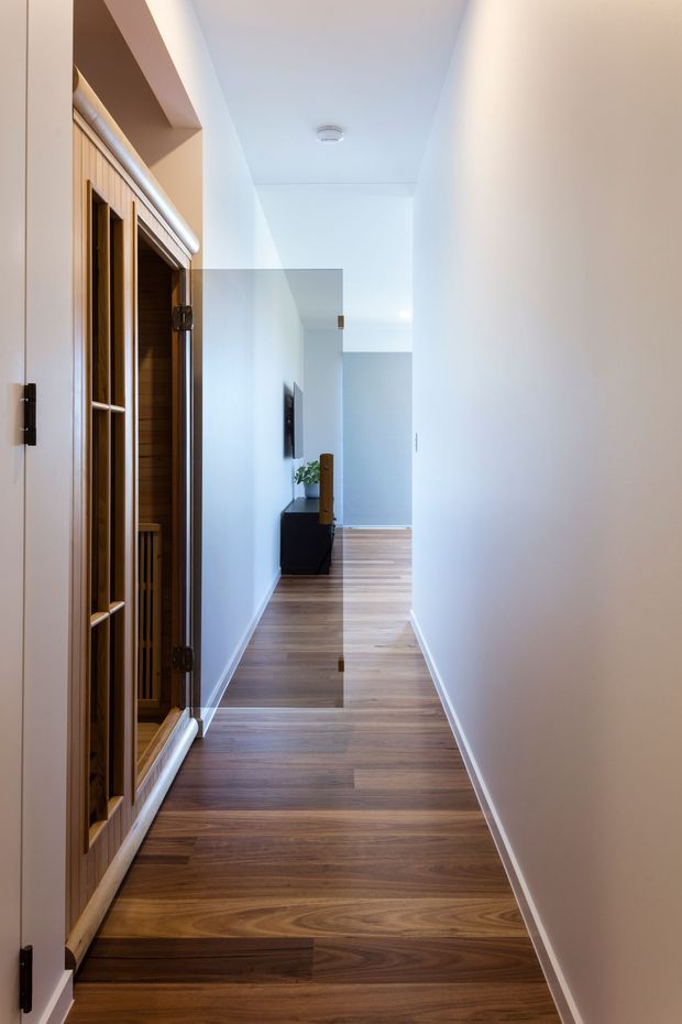 A narrow, bright residential hallway with warm timber flooring and white walls. A timber-framed sauna or steam-room enclosure with glazed doors occupies the left side, while a compact dark console and wall-mounted television are visible farther down the corridor. The space has a clean, contemporary fit-out with simple white trim and recessed circulation.
