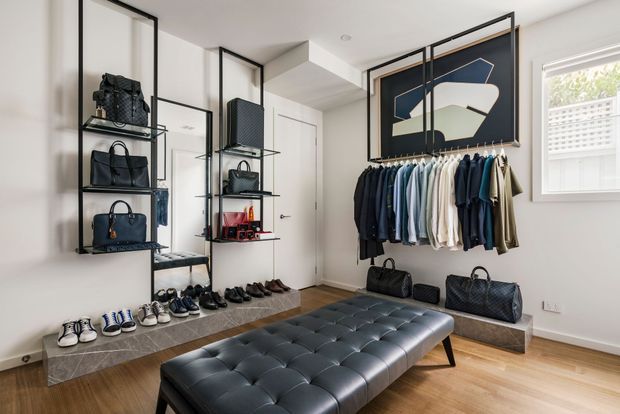 Bright, contemporary walk-in wardrobe or dressing room with open black metal shelving displaying handbags, a full-height mirror, a wall-mounted rail of neatly arranged clothing, shoe storage, and a large black tufted leather bench. The room has pale walls, light timber flooring, a grey tiled raised platform, and a geometric framed artwork.