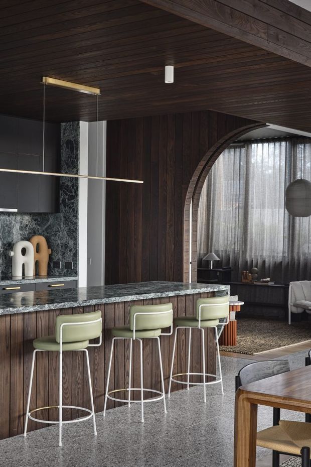 A dark, sophisticated open-plan kitchen and living area. The kitchen features a long timber-clad island with a polished dark stone benchtop, integrated cabinetry, green upholstered bar stools, and a dark marble-look splashback. A timber-lined ceiling and walls create a warm, enveloping atmosphere. Beyond the island is a lounge nook with curved timber arch detailing, sheer curtains, a dark rug, and soft seating. Linear pendant lights and sculptural decor add refined contemporary character.
