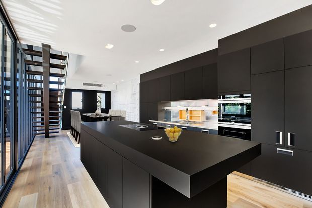 A sleek, contemporary open-plan kitchen with a large black island and matching full-height cabinetry. The island includes an integrated dining overhang, while the rear wall features built-in ovens and a bright preparation area. Light timber flooring, white walls and ceiling, extensive glazing, and a sculptural open-riser staircase create a high-contrast, spacious interior.
