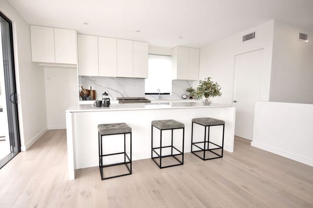 Bright contemporary white kitchen with a large island and three black-framed bar stools. The space features white flat-panel cabinetry, a white marble-look splashback and benchtop, pale timber flooring, integrated appliances, and abundant natural light.