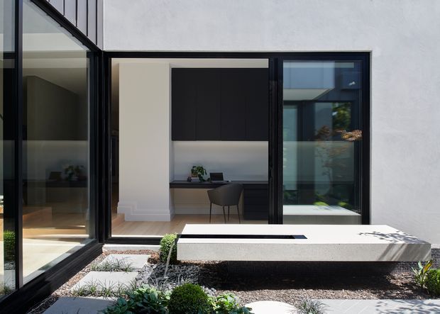 Contemporary courtyard view of a minimalist home office framed by expansive black metal glazing. The built-in desk and dark upper cabinetry are visible through the opening, while a low rectangular water feature or planter sits in the landscaped foreground. White rendered walls, dark gravel, clipped greenery, and strong indoor-outdoor connections define the composition.