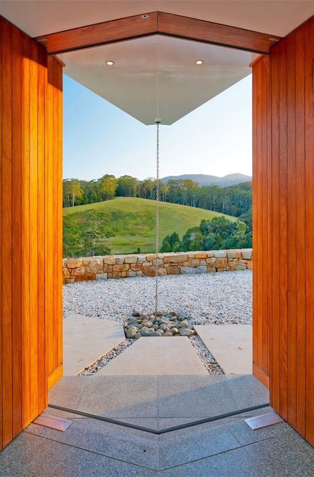 View from inside a timber-lined architectural opening toward a gravel terrace and landscaped rural hills. The space features warm vertical timber wall cladding, a projecting flat soffit with recessed downlights, a hanging chain or rain-chain detail, a low natural-stone retaining wall, and geometric concrete paving with pebble drainage beds. The image emphasizes the connection between the interior threshold and the outdoor landscape.