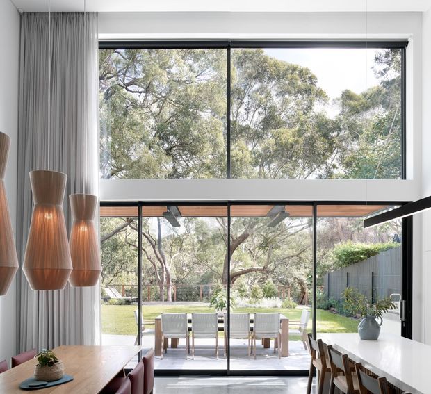 Bright contemporary open-plan dining and kitchen space with a double-height glazed wall opening to a landscaped garden. The room features a large timber dining table, sculptural pendant lights, a white kitchen island with stools, dark-framed glazing, and an outdoor dining area visible beyond the glass.