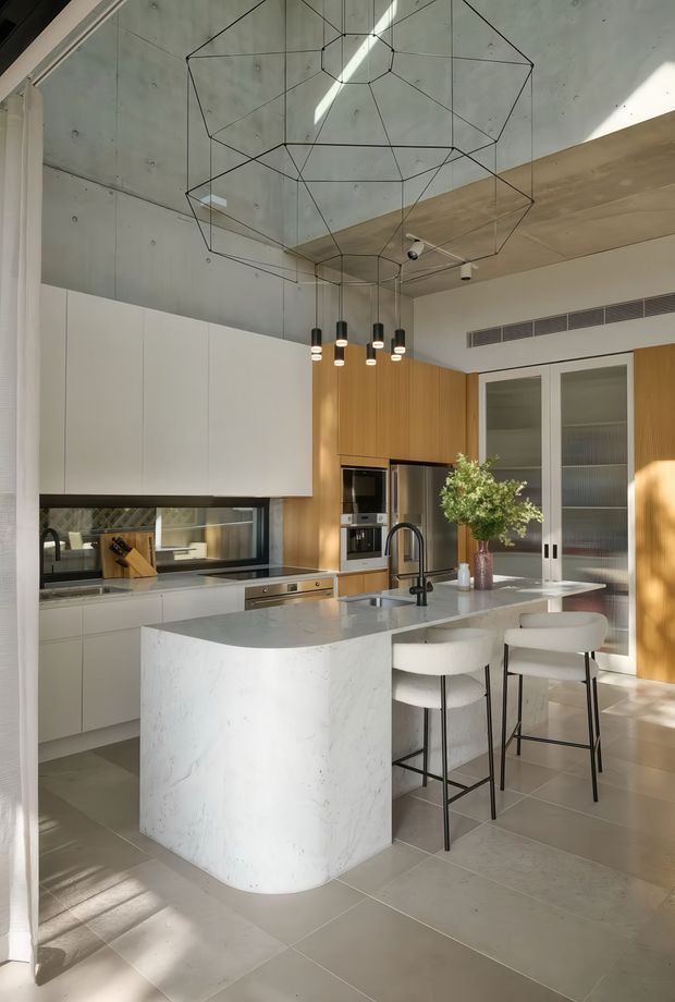 A bright contemporary kitchen with a large white stone island featuring a rounded end and integrated seating for four. The room combines white flat-panel cabinetry, warm timber wall cabinetry, dark-framed internal glazing, and a concrete ceiling with visible joints. Black geometric wire pendant lighting hangs above the island, while integrated appliances, a dark tiled splashback, and tall glazed storage doors complete the refined minimalist fit-out.