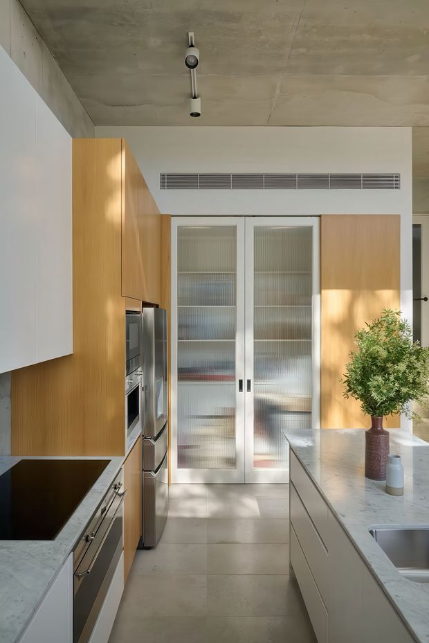 A contemporary galley-style kitchen with pale stone countertops, integrated stainless-steel appliances, light timber wall cabinetry, white lower cabinetry, and full-height ribbed-glass pantry doors. A central island with a marble-like benchtop occupies the right side. The space has a raw exposed concrete ceiling, linear air-conditioning grille, track lighting, and a warm neutral palette.