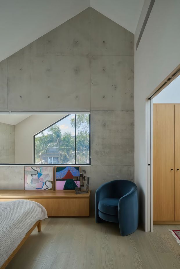 A contemporary bedroom with a dramatic exposed concrete feature wall rising to a peaked ceiling, pale timber flooring, and a built-in timber window seat or low cabinet. A large angular window brings in views of palm trees and a neighbouring building. The room includes a partially visible bed with a light textured cover, a sculptural dark blue armchair, framed artwork, and tall light-timber built-in wardrobe doors. The architecture is minimalist and material-focused, with clean white ceiling planes and strong geometric forms.