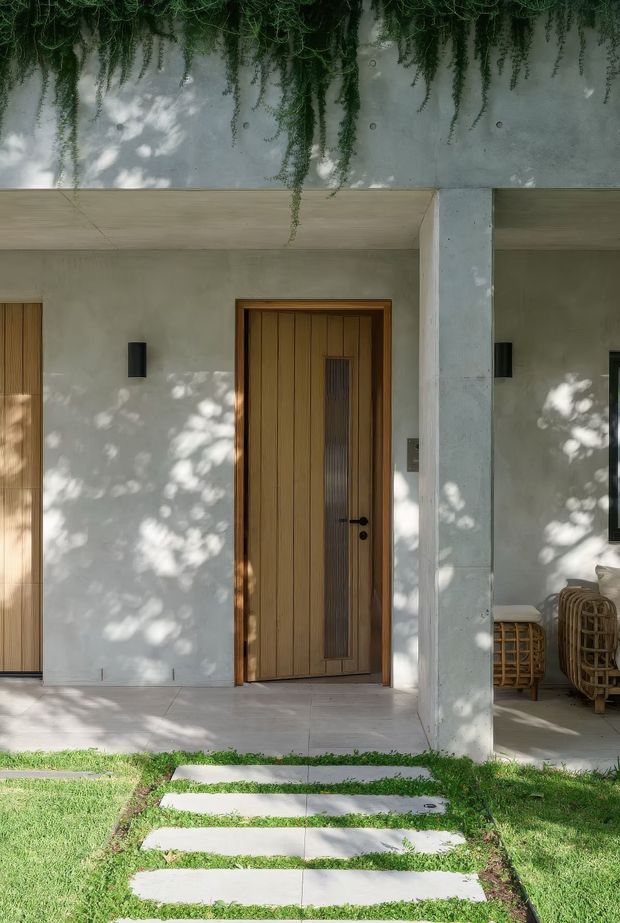 A contemporary residential entry facade viewed from the front garden. The sheltered porch has smooth pale rendered walls, a flat concrete canopy and substantial square concrete column, with warm vertical timber entry doors and dark wall sconces. A stepping-stone path crosses the lawn, while trailing greenery and woven outdoor seating add a natural, relaxed character.