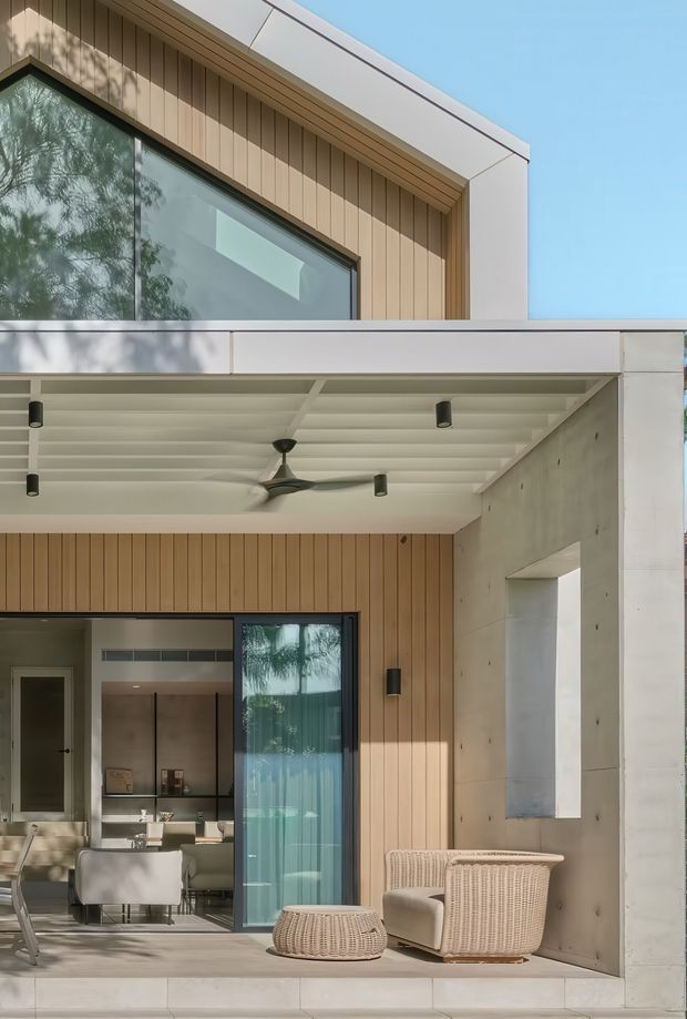 A contemporary residential exterior showing a covered outdoor living terrace beneath an upper-level facade. The architecture combines warm vertical timber cladding, smooth pale concrete walls, large dark-framed glazed openings, and a white slatted soffit with recessed downlights and a ceiling fan. Outdoor wicker lounge furniture sits beside an open connection to a refined dining area visible through the glazing. Bright, warm, clean, and highly detailed presentation with strong indoor-outdoor flow.