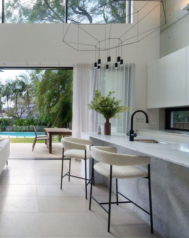 Bright, contemporary open-plan kitchen and dining area with a large white stone island, integrated white cabinetry, black gooseneck tap, and three upholstered bar stools. Full-height glazing and sheer curtains open to a landscaped backyard with a swimming pool. A sculptural geometric pendant light hangs from the high ceiling, and a timber dining table sits beside the island.