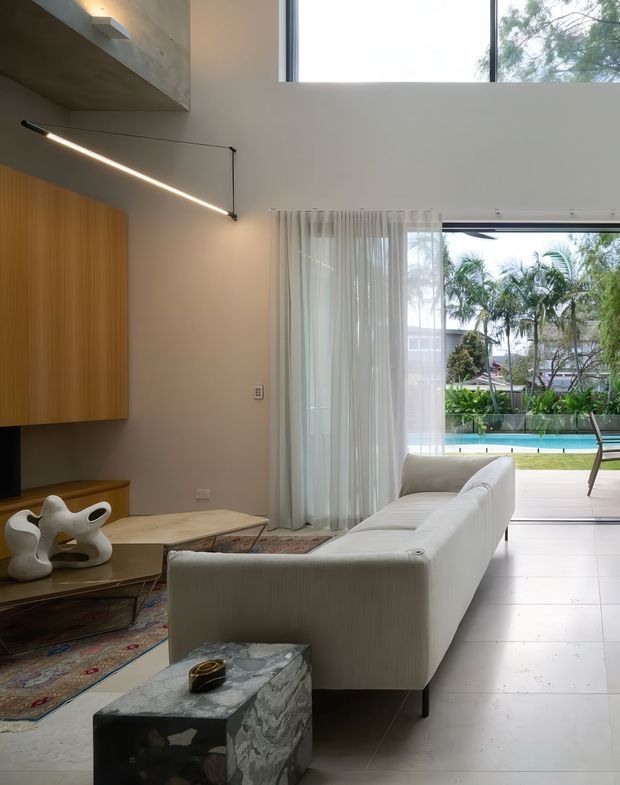 A contemporary double-height living room with a large white sectional sofa, low timber coffee tables, a sculptural side table, patterned area rug, and minimalist linear lighting. Warm wood wall cabinetry contrasts with smooth grey-beige walls and pale tiled flooring. Floor-to-ceiling sliding glass doors and sheer curtains open to a bright pool and tropical garden beyond.