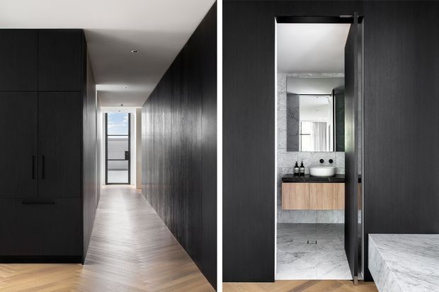 A narrow, refined hallway lined with full-height dark timber cabinetry and panelled walls, leading to a glazed exterior door. A concealed door opens to a compact bathroom with grey stone surfaces, a wall-mounted timber vanity, round basin, mirror, and black fixtures. The foreground also shows the edge of a marble- or stone-topped surface.