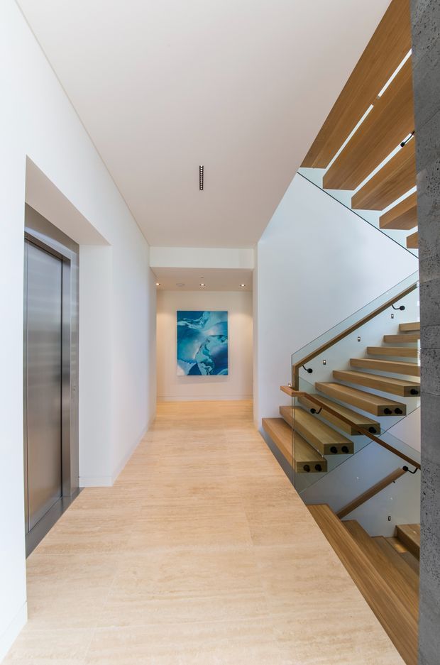 A bright, minimalist interior hallway and stair landing. The space features pale timber flooring, white walls and ceiling, a timber staircase with glass balustrades and slim dark handrails, and a stainless-steel lift door. A blue abstract artwork is displayed at the end of the corridor. Timber slatted ceiling elements and a textured grey structural wall add architectural detail.