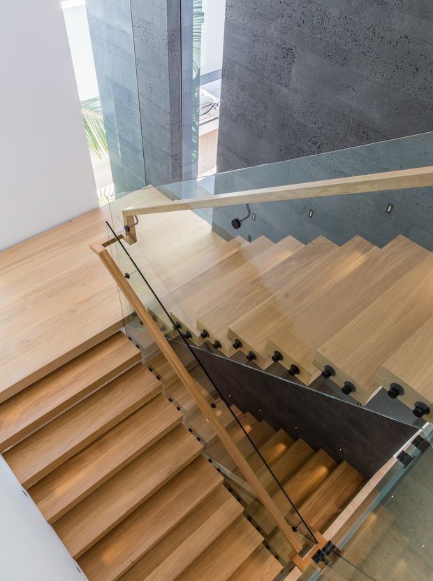 Contemporary interior stairway viewed from above, featuring broad light timber treads, clear glass balustrades with timber handrails and dark metal fittings. A textured dark grey concrete or stone wall forms the stair enclosure, with abundant natural light entering through tall glazing. The multi-level stair composition creates a dramatic open void and strong geometric lines.