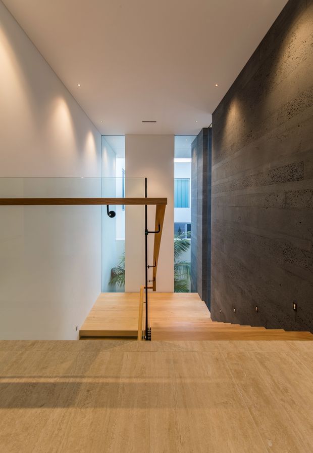 A modern interior stairway viewed from an upper landing, featuring broad light timber steps, a slim black metal handrail with glass balustrade, a tall textured dark concrete feature wall, and warm recessed lighting. Full-height glazing at the stairwell reveals palms and a bright exterior beyond.