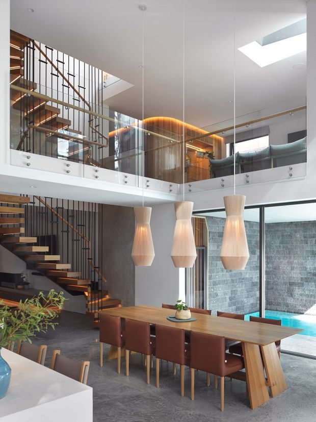 Double-height contemporary dining and living interior with a long timber dining table, brown upholstered chairs, three sculptural pendant lights, an open floating staircase with dark metal balustrade, glass mezzanine balustrades, skylights, and large glazing overlooking a pool. Warm timber and illuminated curved timber detailing contrast with dark stone-like flooring and white surfaces.