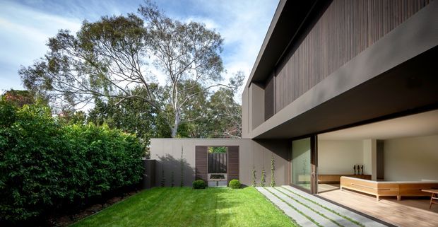 A contemporary two-storey residential courtyard viewed from the landscaped garden. The building has a long cantilevered upper level with dark vertical timber cladding, smooth light-grey rendered walls, extensive glazed openings, and a covered outdoor living area with built-in timber seating. A narrow lawn, clipped hedges, stepping-stone paving, and mature trees create a private garden setting.