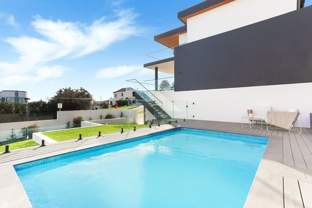 Bright contemporary residential exterior with a large turquoise swimming pool in the foreground, surrounded by pale decking and paving. The home has crisp white rendered walls, a dark charcoal feature facade, projecting flat roof forms, extensive glass balustrades, and an external stairway. Minimal outdoor furniture and manicured landscaping complete the modern poolside setting.