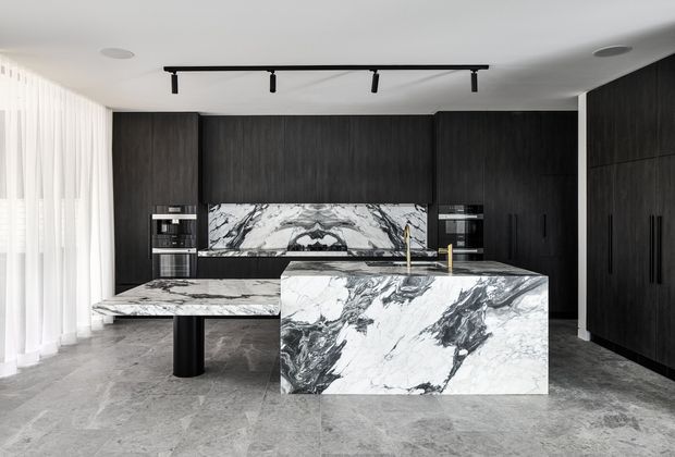 Contemporary kitchen featuring dramatic dark timber cabinetry, a large sculptural marble island with waterfall edge, matching marble benchtops and splashback, integrated appliances, black tapware, and grey stone-look flooring. Sheer floor-to-ceiling curtains and minimalist track lighting contribute to a refined, monochromatic interior.
