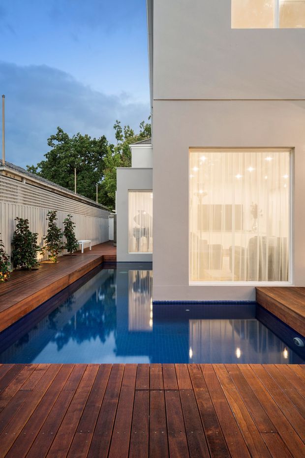 A narrow urban courtyard with a long rectangular swimming pool surrounded by rich dark timber decking. A minimalist pale rendered building forms the right side, with large floor-to-ceiling windows covered by sheer curtains and warm interior lighting. The left boundary has a textured privacy screen and planted greenery, creating a refined private outdoor retreat.