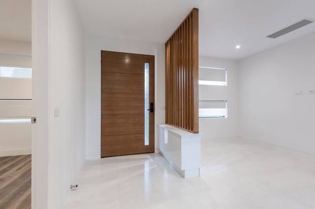 Bright contemporary entry area with a horizontal-grain timber front door, narrow patterned glass sidelight, polished white flooring, and a vertical timber slat screen forming a partial divider around a white built-in ledge. A glimpse of an adjacent room with timber flooring is visible at left, while the main space has recessed lighting, minimalist white walls, and slim horizontal windows.