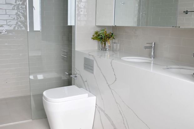 Bright contemporary bathroom featuring a wall-mounted mirrored cabinet, glossy light-grey subway tile walls, a frameless glass shower enclosure, a sleek white toilet, and a long white vanity with integrated basins and prominent grey veining. Chrome tapware and minimal styling create a clean, refined look.