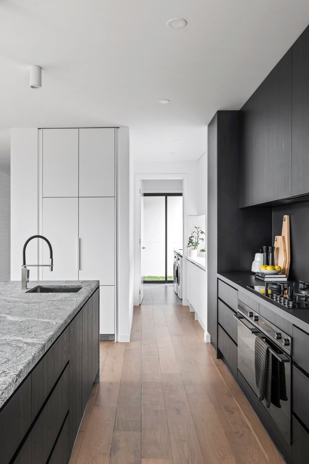 A narrow, contemporary galley kitchen with a large central island, white waterfall-style stone benchtop, integrated sink and black gooseneck tap. The kitchen combines dark timber-look cabinetry and a black splashback wall with tall white storage cabinetry. Integrated cooking appliances line the right side, while pale timber flooring leads toward a bright utility area and exterior doorway. Minimal recessed and cylindrical ceiling lighting create a clean, high-contrast interior.