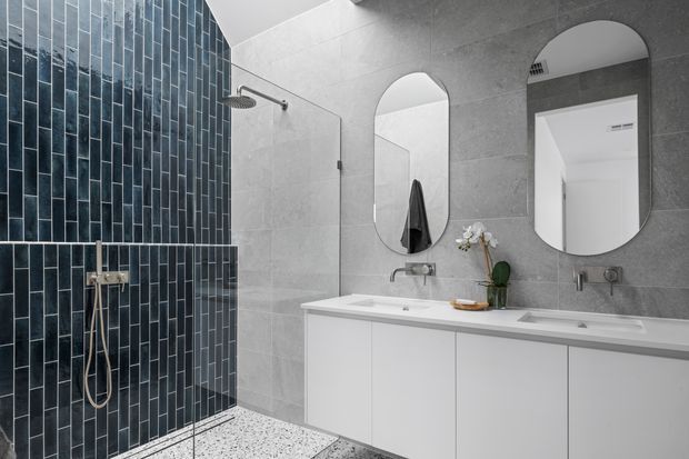 Contemporary bathroom featuring a double vanity with white cabinetry, large rounded-arch mirrors, grey wall tiling, a dark blue-grey vertical tile shower feature wall, frameless glass shower screen, and terrazzo-style flooring. The space is clean, refined, and moderately bright with strong tonal contrast between the dark shower tiles and light vanity.
