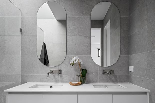 Contemporary double-vanity bathroom with large-format grey wall tiles, a white built-in vanity with two integrated basins, brushed metal wall-mounted taps, and paired arched mirrors. A small orchid and timber accessory add a restrained natural accent.